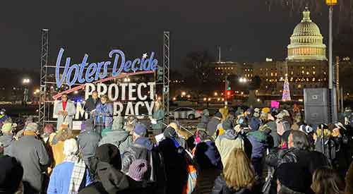 People gathered at outdoor event with Voters Decide Protect Democracy sign and U.S. Capitol building in background